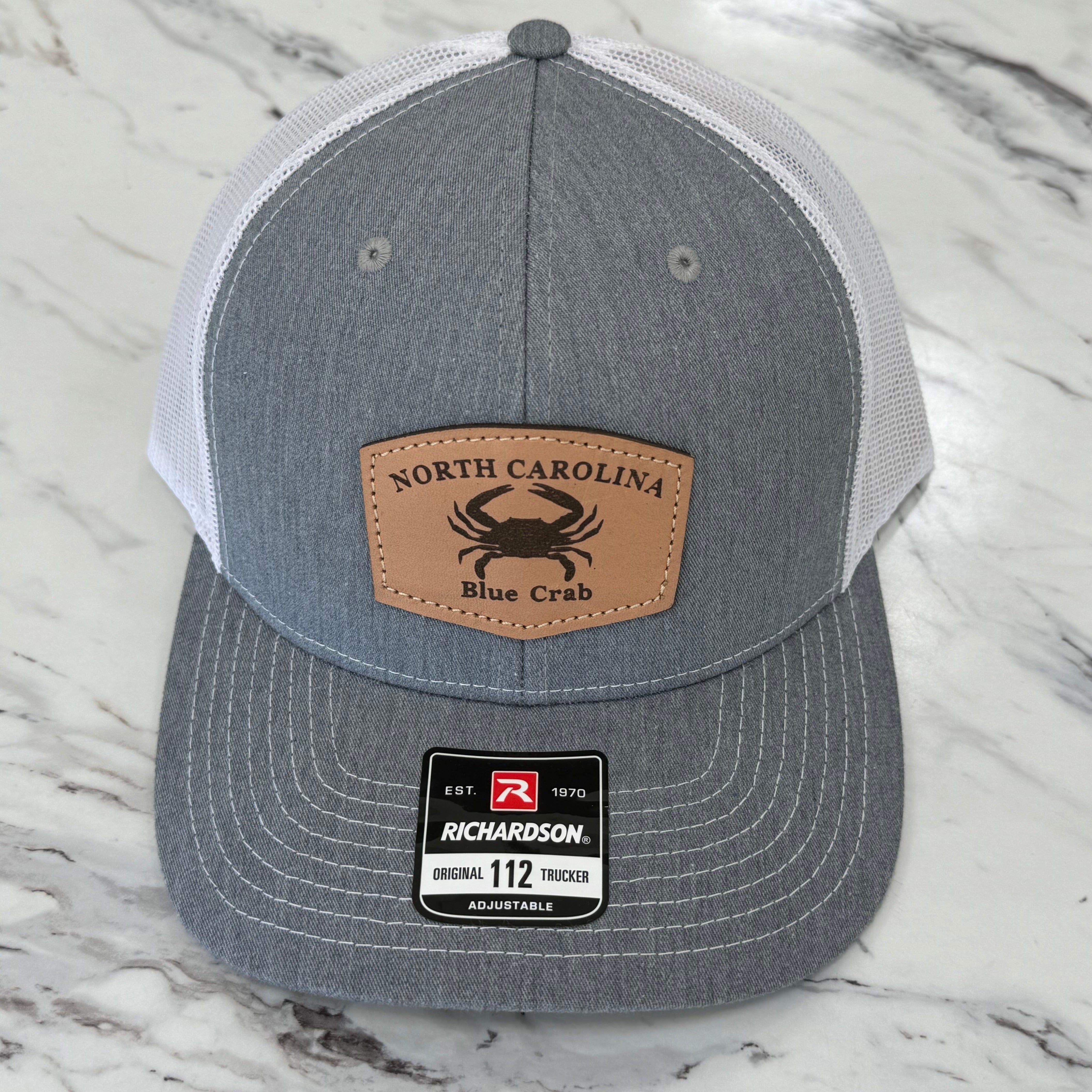 White and grey hat with leather emblem that has a blue crab and North Carolina written on it