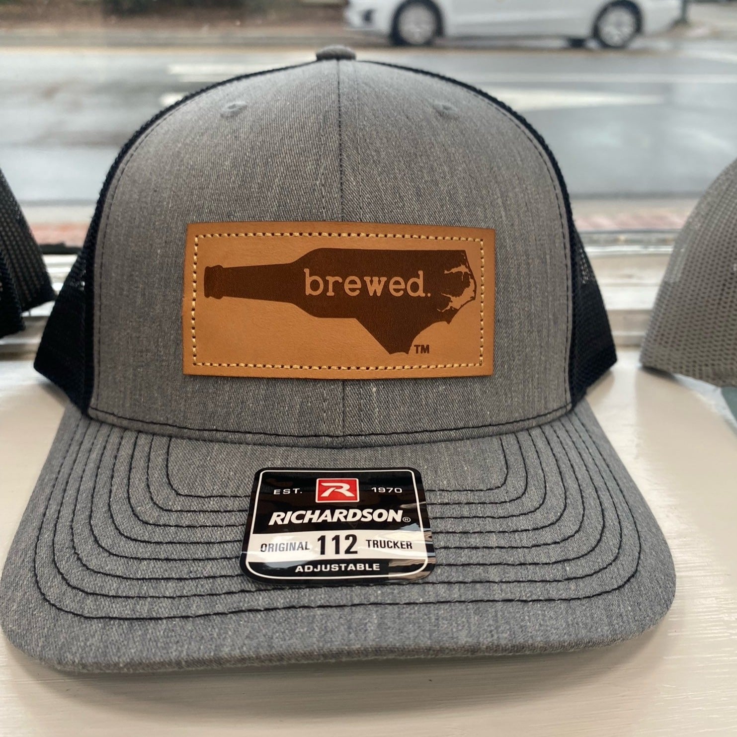 Black and heathered grey hat with a leather patch that says "Brewed" inside the shape of North Carolina. 
