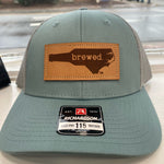 Load image into Gallery viewer, Blue and grey hat with a leather patch that says &quot;Brewed&quot; inside the shape of North Carolina. 
