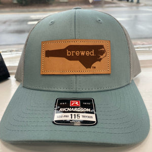 Blue and grey hat with a leather patch that says "Brewed" inside the shape of North Carolina. 