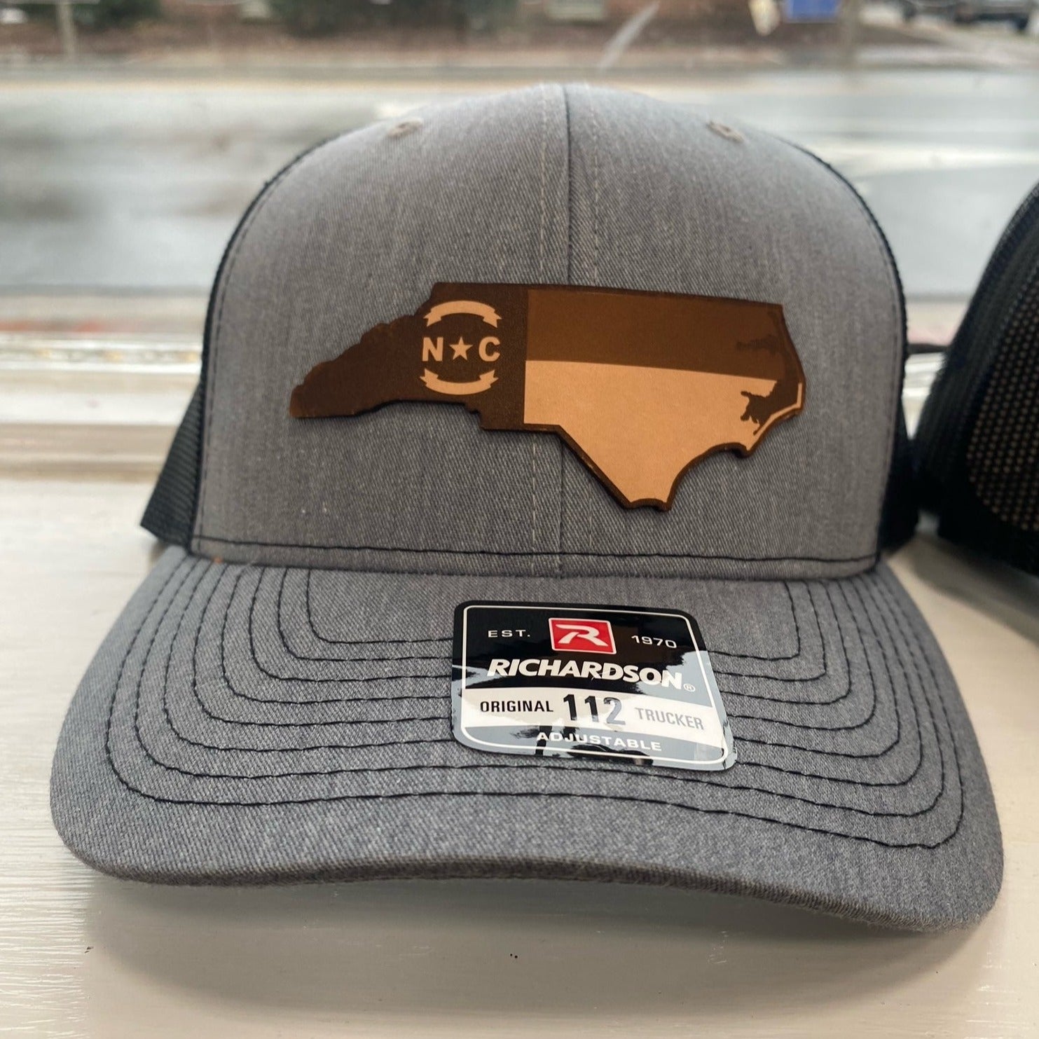 Grey and black hat with a North Carolina shape emblem with the NC flag on it